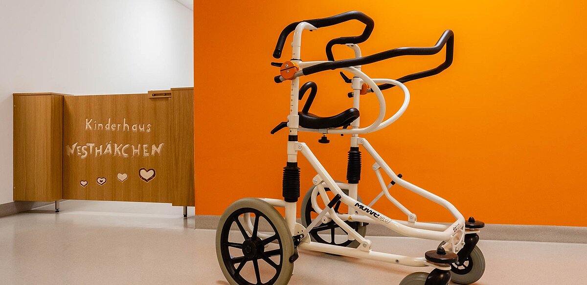 Das Bild zeigt eine Gehhilfe und im Hintergrund eine hüfthohe Schwingtür. Auf der Schwingtür steht "Kinderhaus Nesthäkchen"