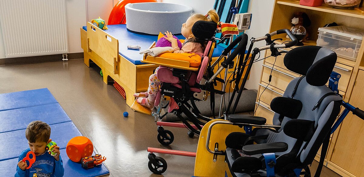 Aufnahme von 2 Kindern. Eines beim Spielen auf dem Boden. Das andere Kind im Rollstuhl sieht aus dem Fenster. 